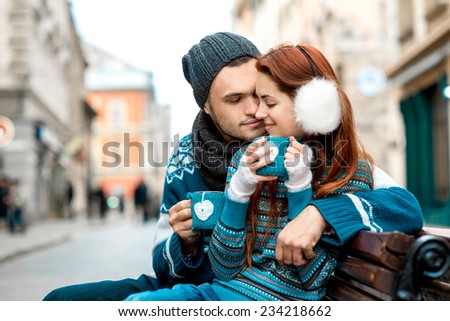 Young loving couple dressed in blue sweater sitting with knitted coffee cups on the bench at the old city in winter