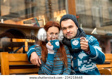 Young loving couple dressed in blue sweater sitting with knitted coffee cups on the bench near cafe in winter