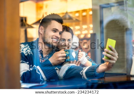 Young loving couple dressed in blue sweater sitting with knitted coffee cups and making selfie photo at the cafe in winter