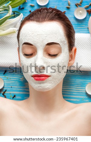Young woman with spa facial mask on her face lying on blue table with flower, candles and sea salt in the beaty salon