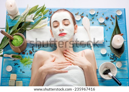 Young woman with spa facial mask on her face lying on blue table with flower, candles and sea salt in the beaty salon