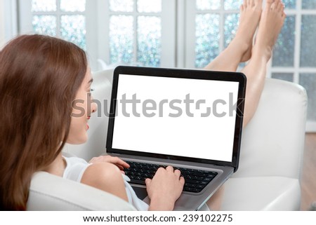 Smiling young woman using laptop with empty screen lying on the couch at home