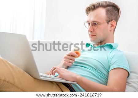 Handsome man eating apple and working with laptop laying on the couch at home.