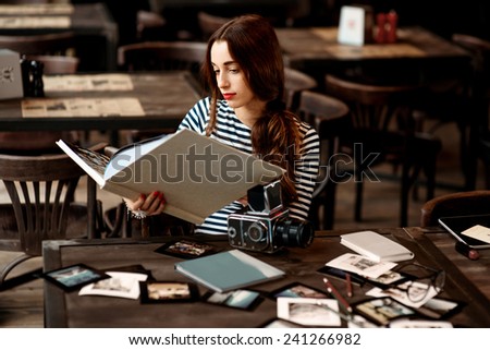 Young woman photographer watching photo album with old 6x6 frame camera and printed photos on the table sitting in the cafe