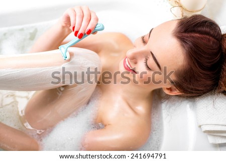 Young and positive woman shaving her legs in the bath with foam and candles.