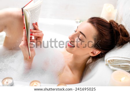 Young woman reading book while lying in the bath with foam and candles