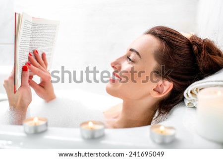 Young woman reading book while lying in the bath with foam and candles