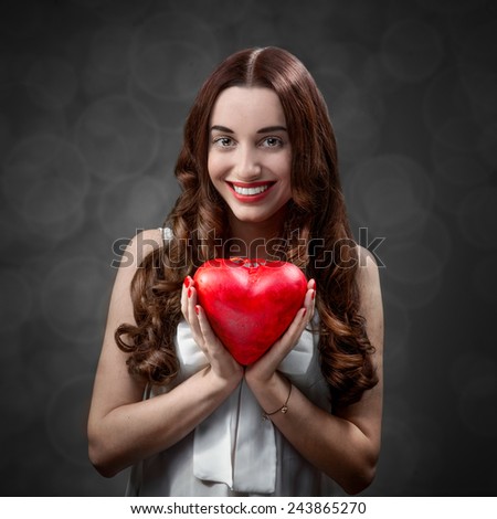 Happy and joyful young woman holding red heart on grey background in studio. Happy valentines greeting concept