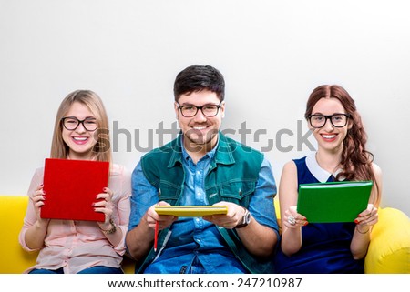Three young students or nerds holding colorful books and sitting on the yellow couch