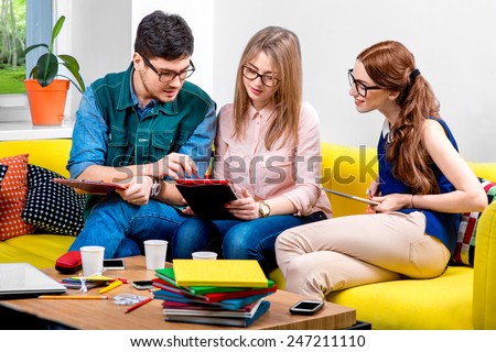 Three young friends or students working with digital gadgets on the yellow couch at home