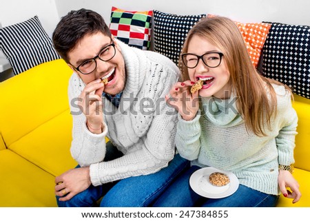 Young couple in sweater and eueglasses eating cakes on the couch at home