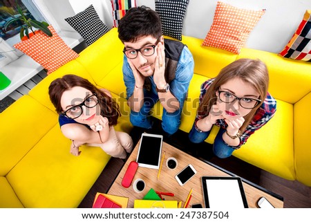 Three nerds in eyeglasses sitting on the couch and looking at camera with different gadgets on the background