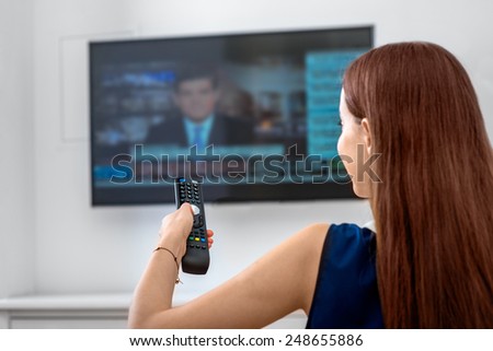 Young woman watching news on television with remote sitting on the sofa at home. Back view