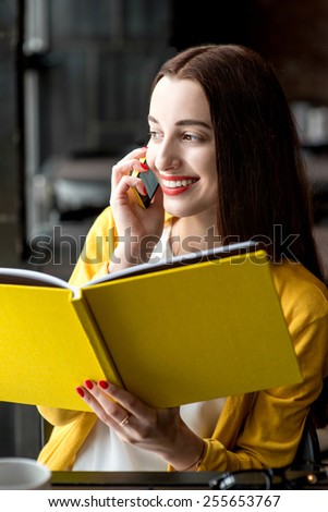 Young smiling woman talking with phone while holding yellow book in the dark interior