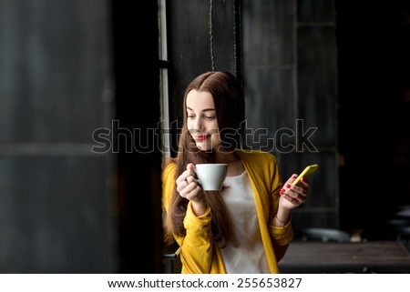 Young and pretty woman in yellow sweater using phone holding a cup of coffee in the dark cafe interior