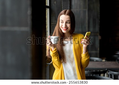 Young and pretty woman in yellow sweater using phone holding a cup of coffee in the dark cafe interior