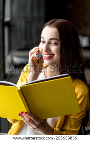Young smiling woman talking with phone while holding yellow book in the dark interior