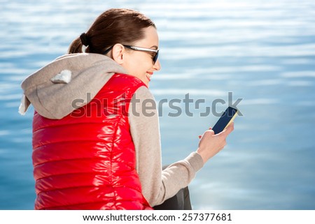 Sport woman dressed in warm clothes resting with phone on the promenade near the water in Spring