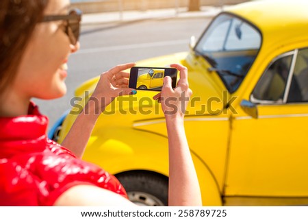 Young woman photographing with smart phone old yellow car on the street