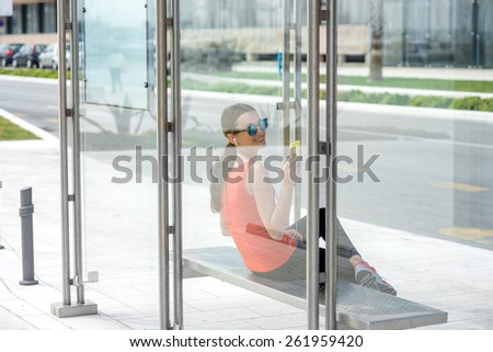 Young sport woman with phone waiting at the bus station. View through the glass reflection