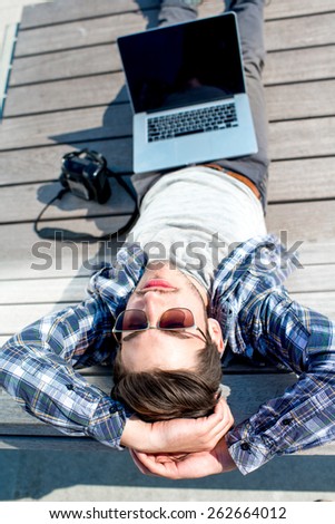 Young photographer in casual clothes resting with laptop and camera on the wooden sunbed. Top view