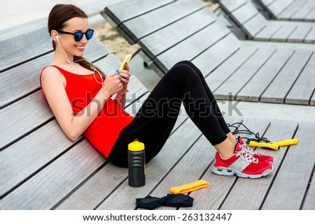 Young sporty woman listening to the music on the wooden sunbed with jump rope, gloves and drink bottle on background