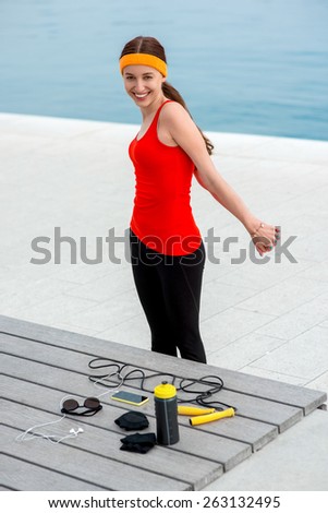 Young sporty woman having exercise on the promenade near the sunbed with jump rope, phone, gloves and drink bottle