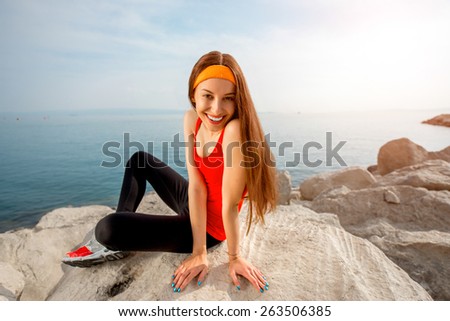 Young sport woman in red shirt and sneakers sitting on the rocky beach in the morning
