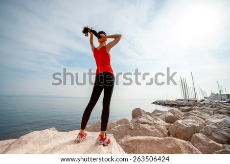 Young sport woman in red shirt and sneakers standing on the rocky beach and looking at the sun in the morning. General plan