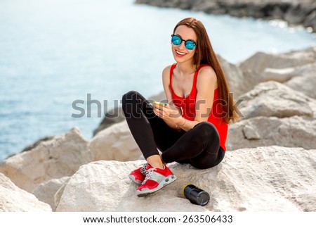 Young sport woman in red shirt and sneakers having rest with mobile phone on the rocky beach in the morning