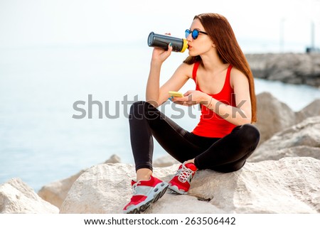 Young sport woman in red shirt and sneakers drinking water on the rocky beach in the morning