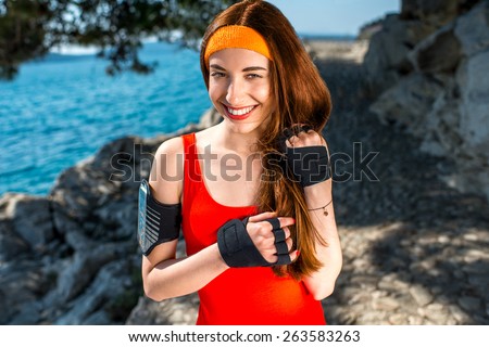 Sport woman in red shirt with sports gloves and phone holder looking at camera in the park near the sea