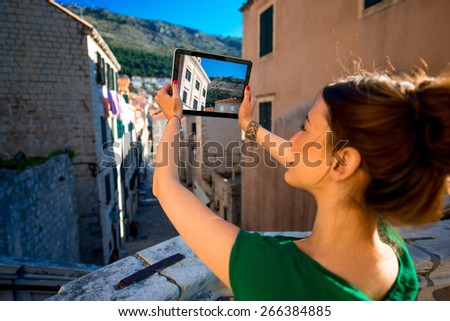 Young woman photographing with digital gadget old buildings in Dubrovnik old city center. Back view with tablet screen.