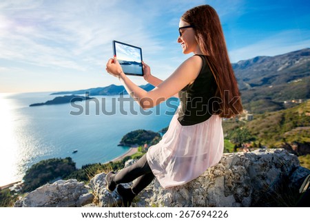 Young woman in dress photographing with digital table sea scape sitting on the mountain top