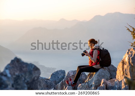 Young traveler photographer with photo camera and backpack sitting on the top of mountain and looking on the sunset