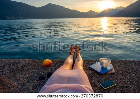 Woman\'s legs on the pier with apple, coffee cup and books at beautiful sunset