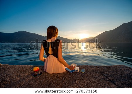 Young woman sitting with coffee cup on the pier at sunrise with beautiful mountains and sea on background