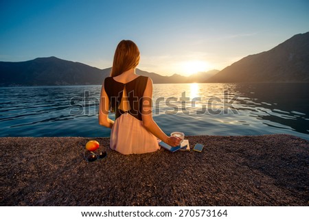 Young woman sitting with coffee cup on the pier at sunrise with beautiful mountains and sea on background