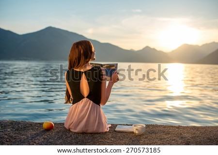 Young woman reading book and looking at beautiful sunrise on the pier with sea and mountains on background