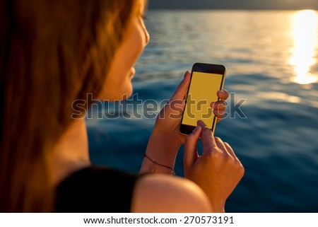 Young woman using phone with empty screen near the sea at sunrise.