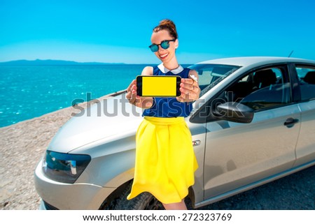 Young and cute woman showing phone screen standing near the car. Navigation or travel phone program concept.