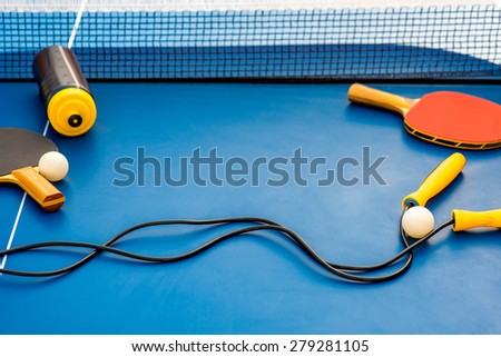Table tennis rackets, balls, rope and drink bottle on a blue table with net and space for text