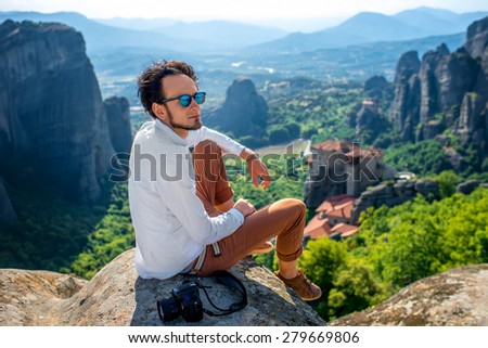 Professional well-dressed photographer sitting on the top of mountain on beautiful scenic clif background near Meteora monasteries in Greece