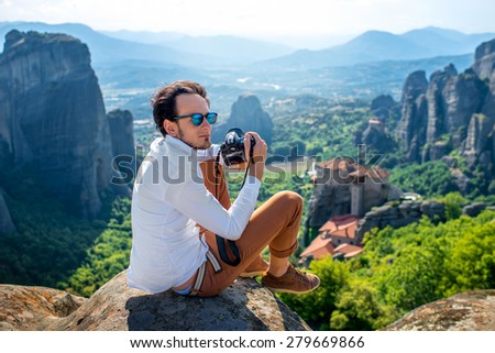 Professional well-dressed photographer sitting on the top of mountain on beautiful scenic clif background near Meteora monasteries in Greece