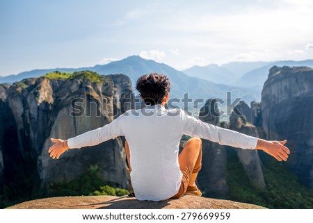 Well-dressed man sitting on the rocky mountain on beautiful scenic clif background near Meteora monasteries in Greece. Back view.