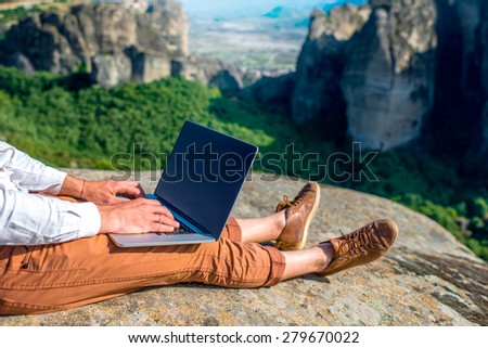 Well-dressed man working with laptop sitting on the rocky mountain on beautiful scenic clif background. Close up view with no face focused on laptop with hands