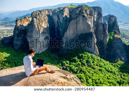 Well-dressed man working with laptop sitting on the rocky mountain on beautiful scenic clif background near Meteora monasteries in Greece. Back view, general plan.