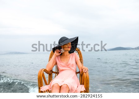 Young woman in pink dress and big black hat resting in the wicker chair on the beach in cloudy weather.