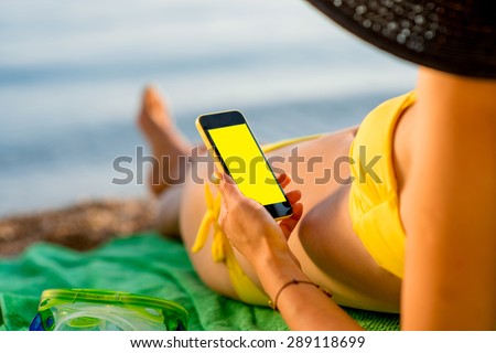 Young woman in swimsuit using mobile phone with empty screen for copy paste lying on the green towel on the beach. Focused on the hand with phone.