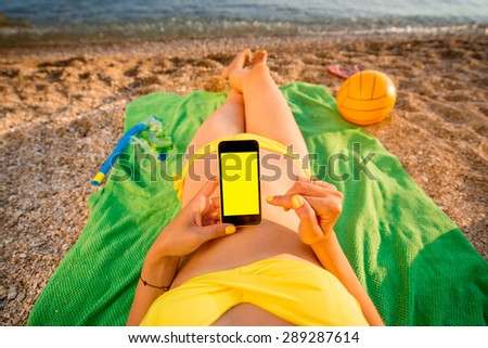 Young woman in swimsuit using mobile phone with empty screen for copy paste lying on the green towel on the beach. Top view focused on the hand with phone.